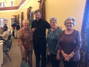 ladies in hats and gloves with Fr. Kurtis Wiedenfeld at Centennial Tea